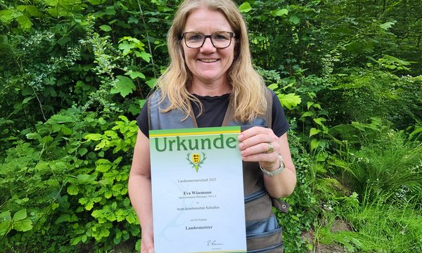 Eine Frau hält eine Urkunde mit der Aufschrift: "Landesmeisterschaft 2025, Eva Wiezmann, 354 Punkte, Landesmeister" in einem üppig grünen Wald.